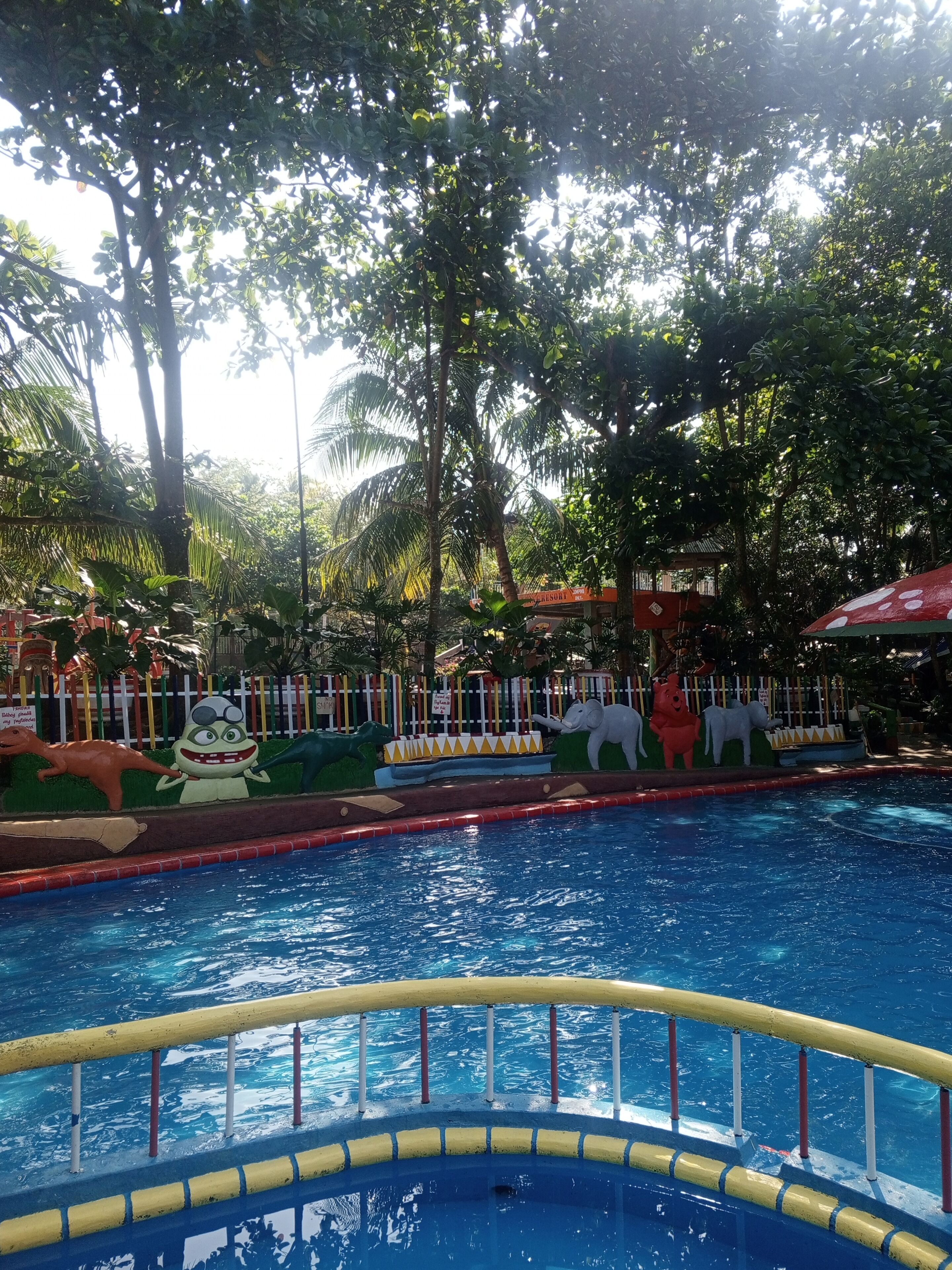 A swimming pool with a yellow and red poolside barrier, surrounded by lush greenery and various colorful animal sculptures.