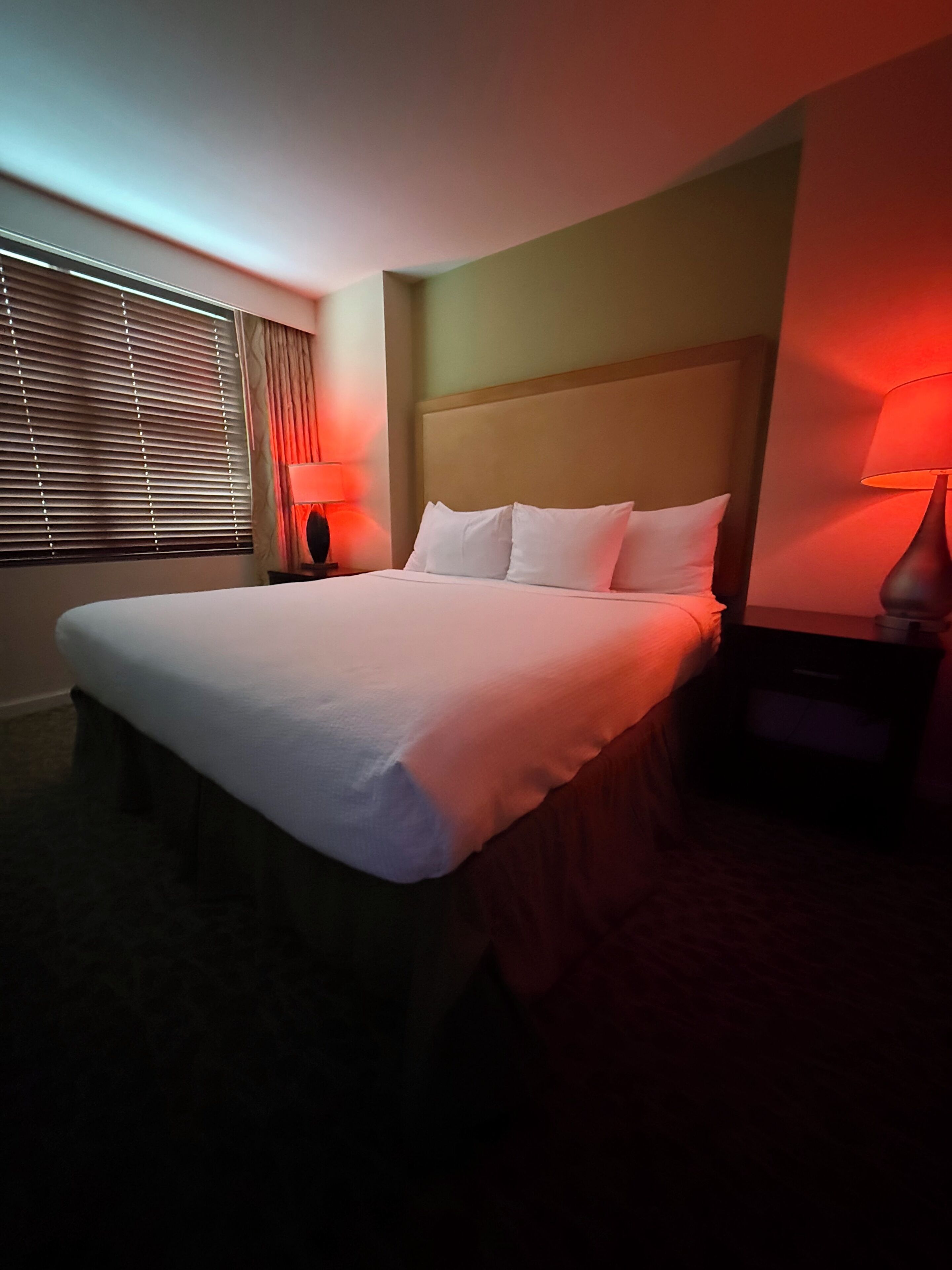 Quarto de hotel com cama, duas luminárias de cabeceira, janela com persianas e cabeceira.