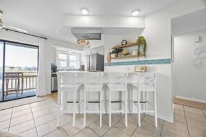 A modern kitchen with white bar stools, a large window, and a view of the city. - New! Coastal Condo - Pool with Marsh + Ocean Views (Carolina Beach)