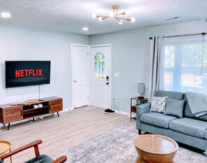 A living room with a flat-screen TV displaying 'NETFLIX,' a wooden entertainment unit, a grey sofa, and a patterned rug. - Indy-Themed Classic: Ode to Hoosier Spirit (Indianapolis)