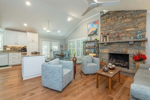 A spacious living area with a stone fireplace, a kitchen with white cabinets, and a dining area with blue chairs. - Cherry On Top | Modern Montreat Home with Screened Porch & Partial Mountain Views! (Montreat)
