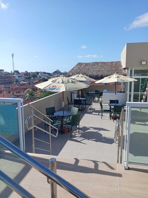 Eine Dachterrasse mit Sitzgelegenheiten im Freien, Blick auf die Stadt und ein Gebäude mit einem Reetdach.