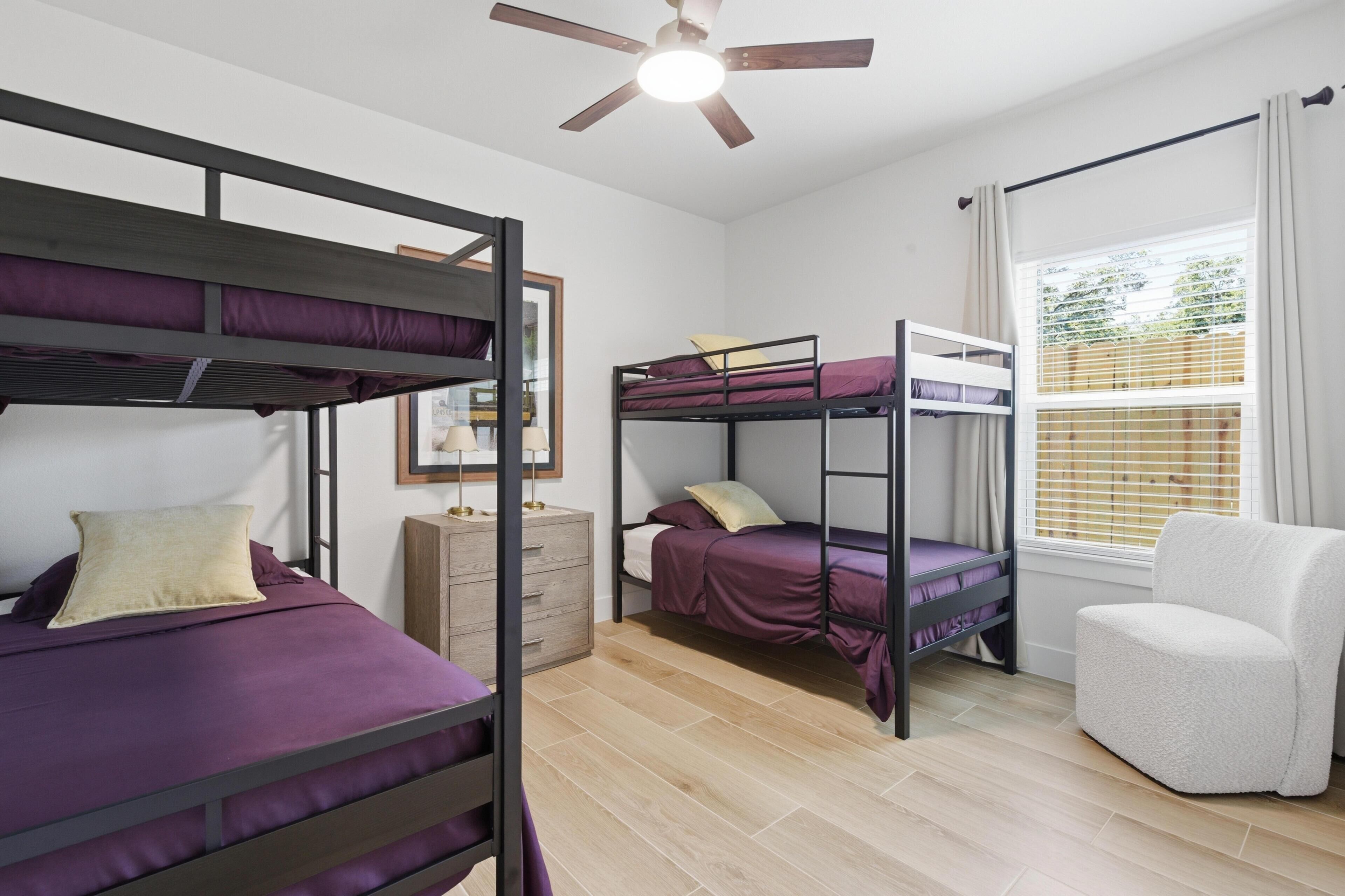 A dormitory room with bunk beds, a ceiling fan, a window with blinds, a chair, and a dresser.