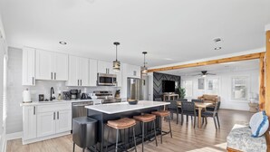 A modern kitchen with white cabinets, a central island with stools, a dining area with a table and chairs, and a living space with a sofa and wooden beams. - Bijou House Unit 2 - 5BR w/ Ocean Views + Heated Pool & Hot Tub (Walk to Beach!) (North Myrtle Beach)