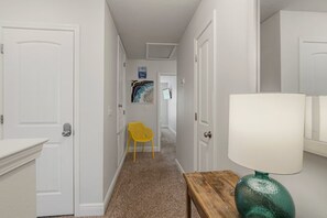A hallway with white doors, a yellow chair, a wooden table with a green lamp, and a framed picture on the wall. - Modern Townhouse. Beach Gear Included! Near Publix Sports Park! (Panama City Beach)