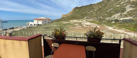 A balcony with a table and chairs, overlooking a beach and a white building with a red roof.