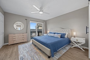 Une chambre à coucher avec un lit, un ventilateur de plafond, une commode, un miroir et une vue sur l’extérieur, accessible par une porte coulissante.