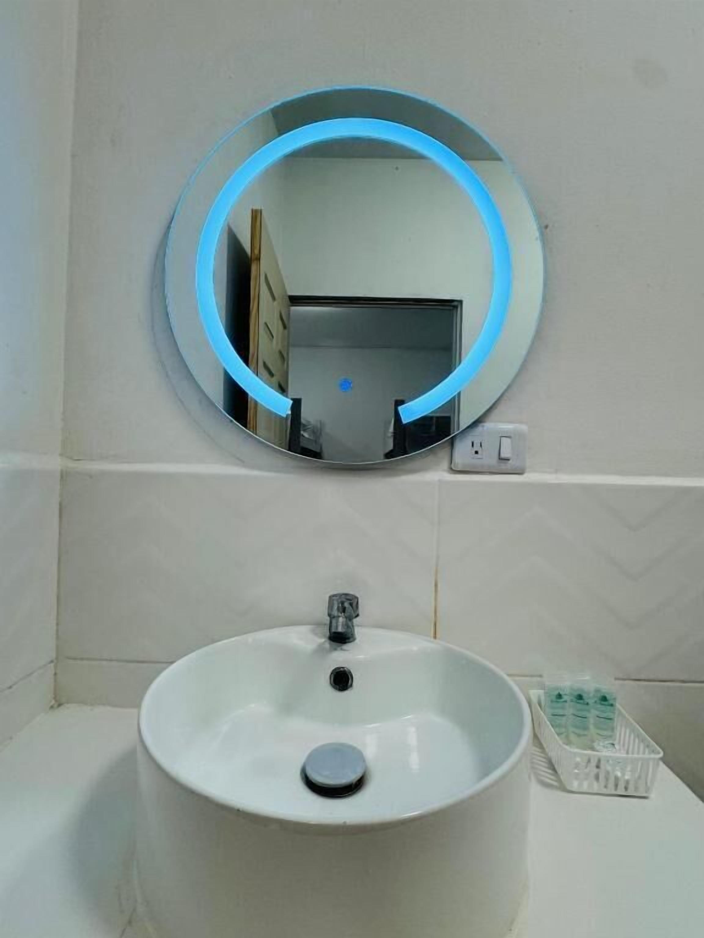 Un baño con un espejo redondo, un lavamanos blanco y una pequeña cesta de artículos de aseo.