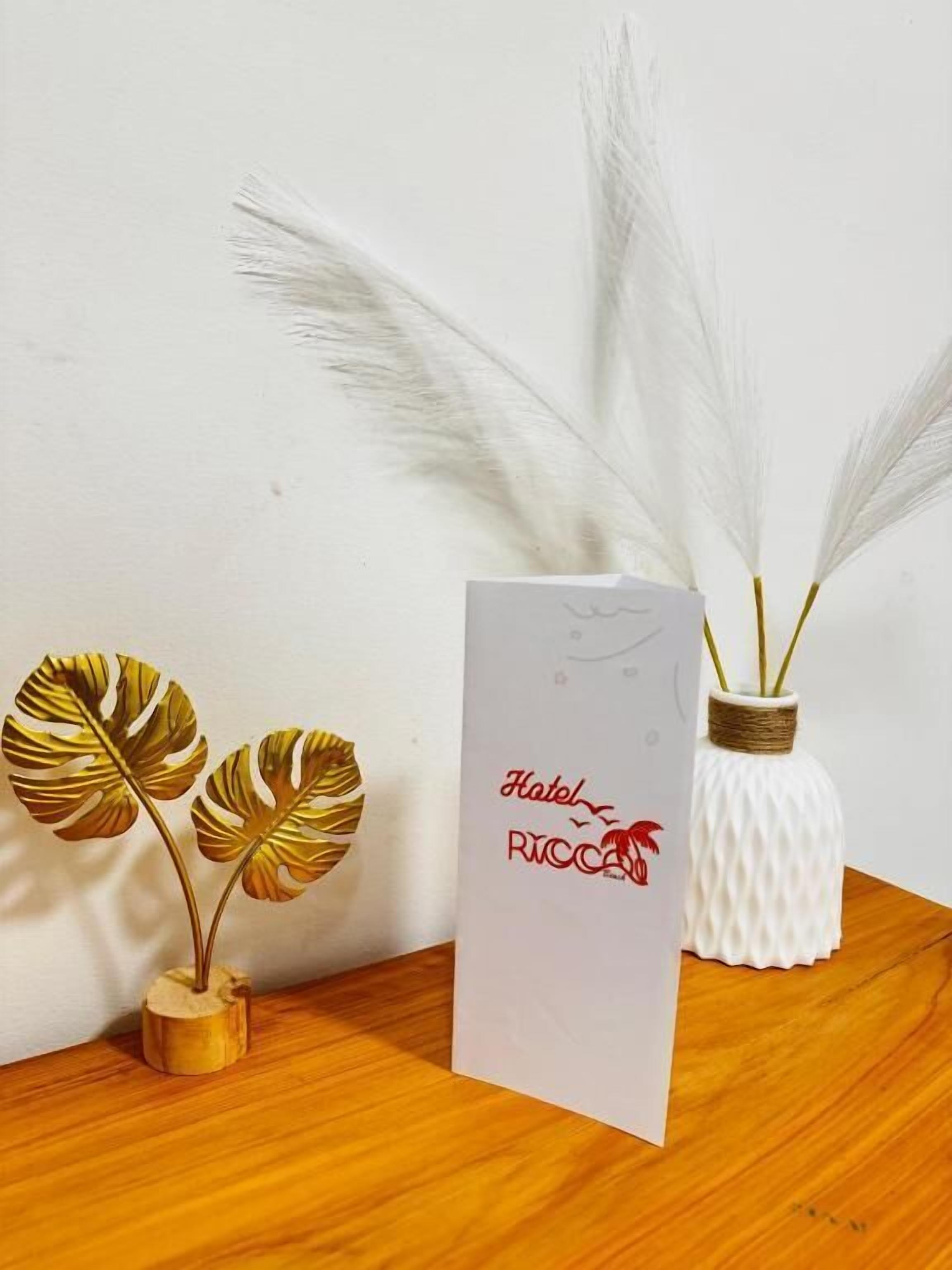 Een wit bord met de tekst 'Hotel Ricca' en een logo, een witte vaas met gedroogd pampa gras, en twee gouden kunstbladeren in een houten vaas.