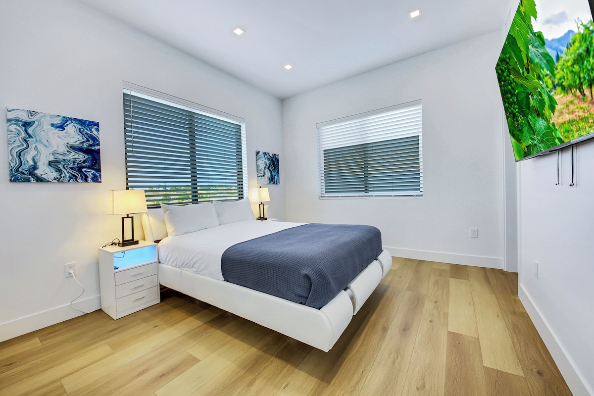 Ein modernes Schlafzimmer mit Bett, Nachttischen, Wandbildern und einem großen Fenster.