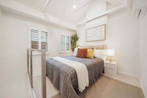 Ein Schlafzimmer mit einem Bett, Nachttischen, einem Fenster mit Rollläden, einem Wandbild und einer Pflanze.