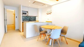 A modern kitchen with a microwave, a herringbone backsplash, and a small island with a sink. There is a dining area with a round table and four chairs. - Apartamento Blue Paradise Punta Romana by VACAY L'Escala (L'ESCALA)