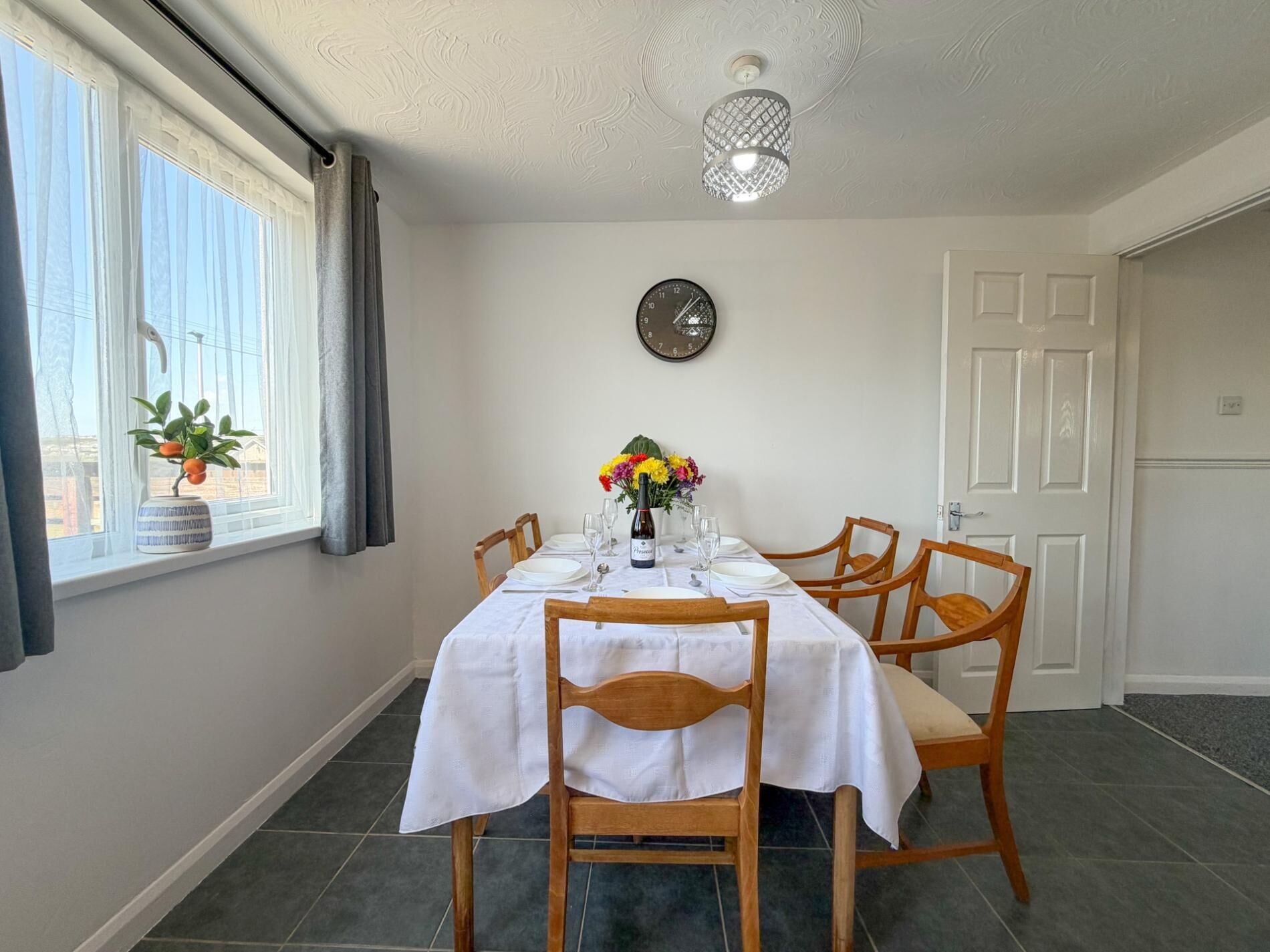 A dining area with a table set for four, a potted plant by the window, a wall clock, and a door.