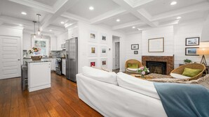 Uma sala de estar moderna com um sofá branco, uma lareira e uma cozinha ao fundo.