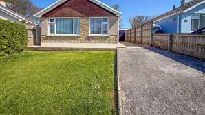 A single-story house with a stone facade, a pitched roof, and a front yard with a lawn and a gravel driveway. - Indigo Blue - 2 Bedroom Holiday Home - Manorbier (Tenby)