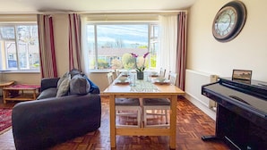 A living room with a sofa, dining table, and piano. - Indigo Blue - 2 Bedroom Holiday Home - Manorbier (Tenby)