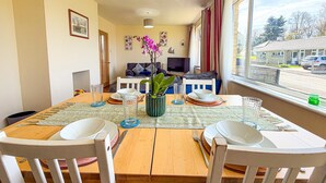 A dining area with a wooden table set for four, a vase with flowers, and a view of the street through large windows. - Indigo Blue - 2 Bedroom Holiday Home - Manorbier (Tenby)