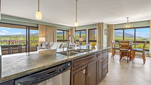 A kitchen with a large island, a dining area with chairs, and a view of the landscape. - Remodeled Corner 2BR with Mountain and Golf Course Views (Lahaina)