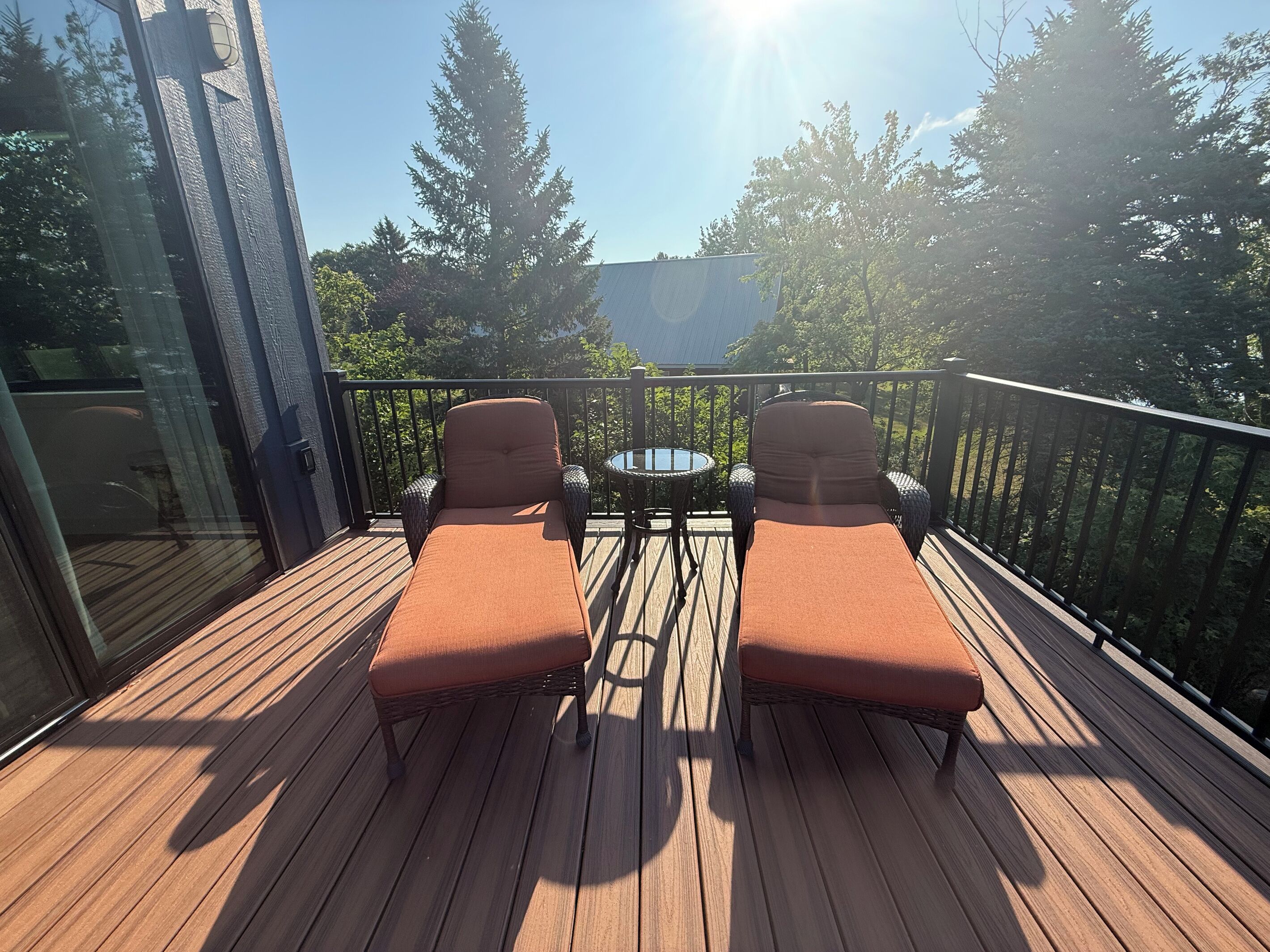 A wooden deck with two lounge chairs and a small table, overlooking a forested area.