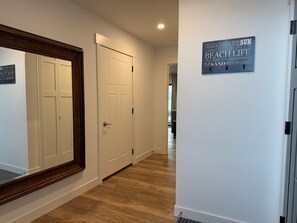A hallway with wooden flooring, white walls, and a large mirror. There is a door with a sign on the wall.