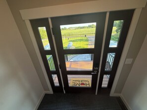 Vista a través de una puerta con amplios ventanales, mostrando una terraza de madera y un campo de césped.