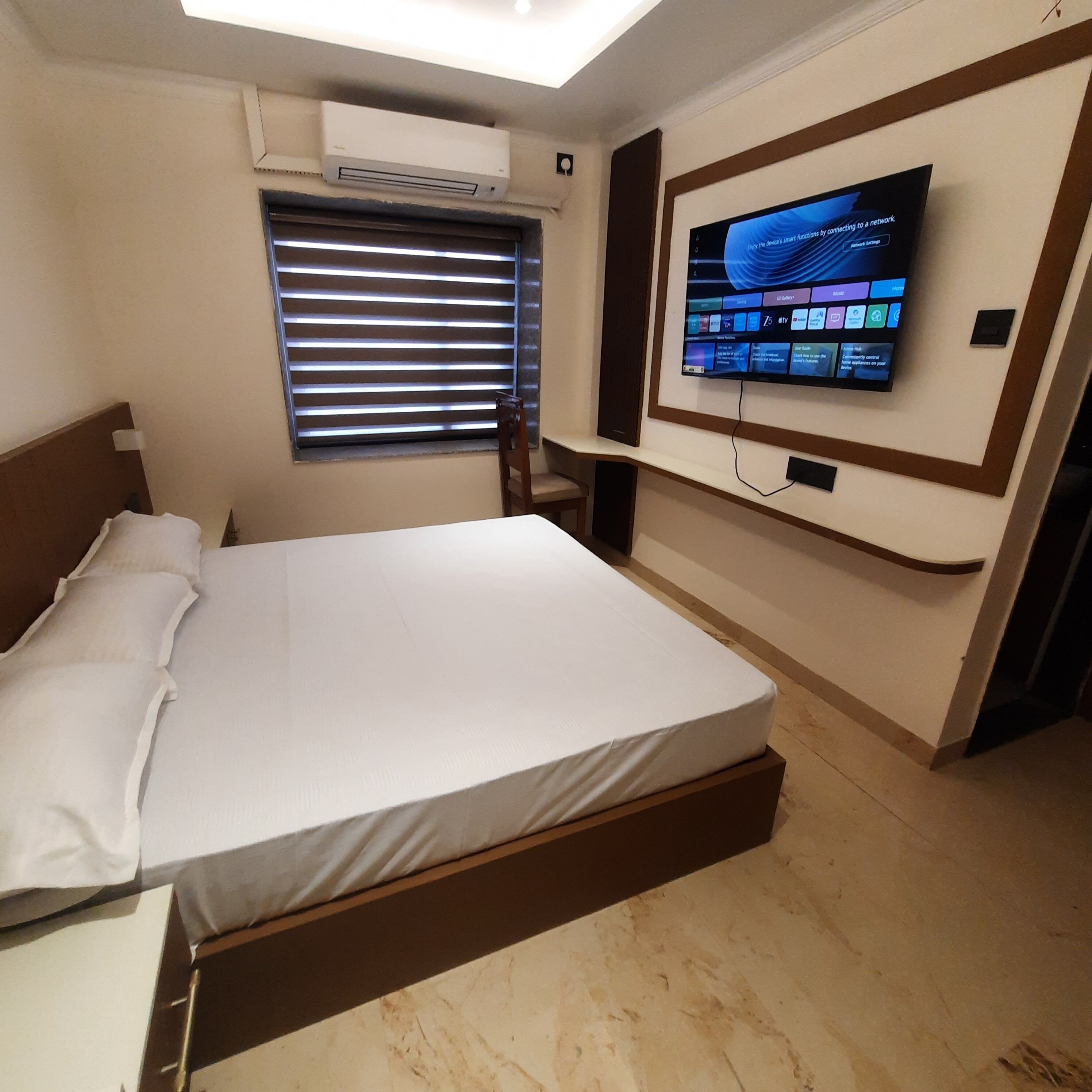 Habitación de hotel con cama, televisión montada en la pared, ventana con persianas y aire acondicionado.