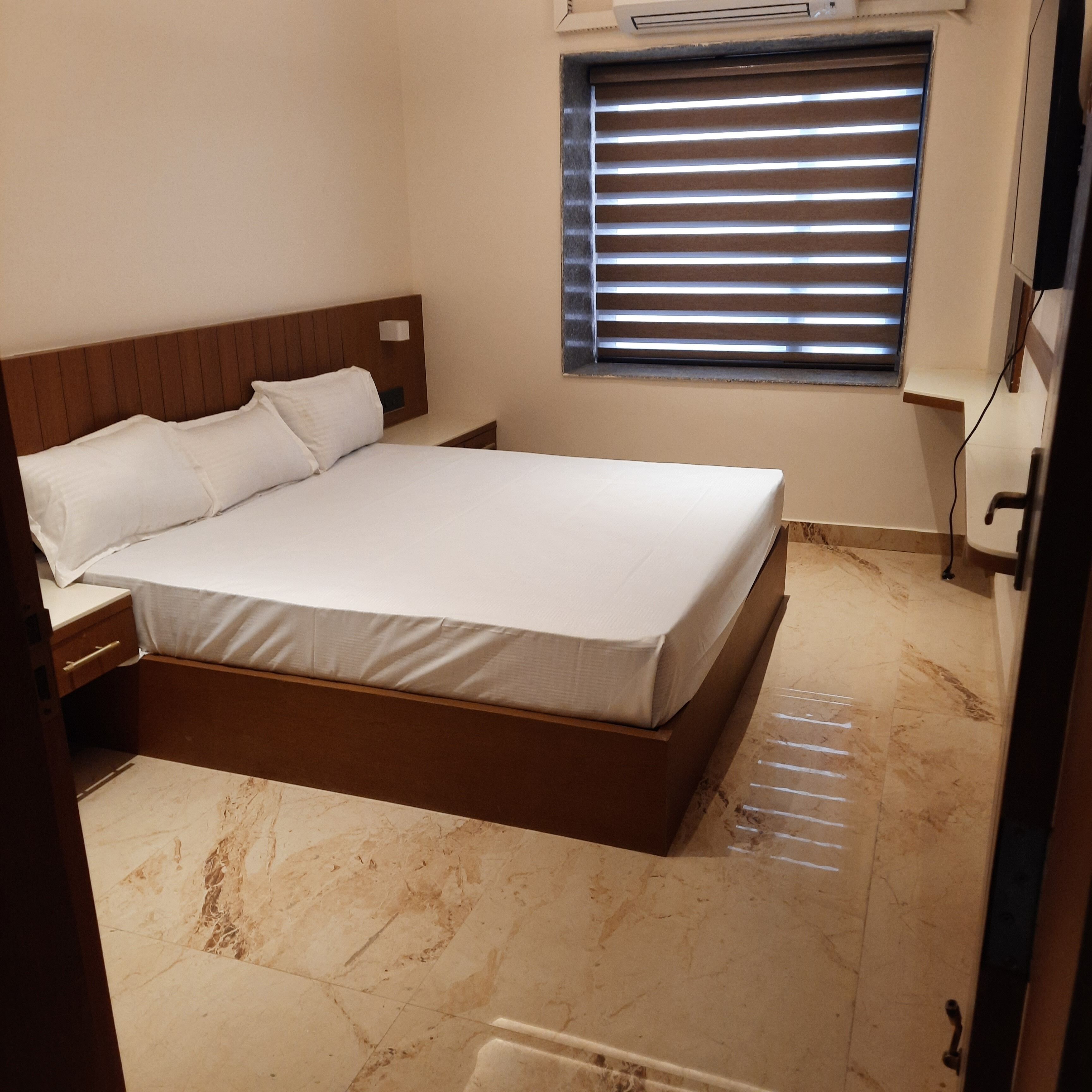 A bedroom with a bed, a nightstand, a window with blinds, and an air conditioning unit.