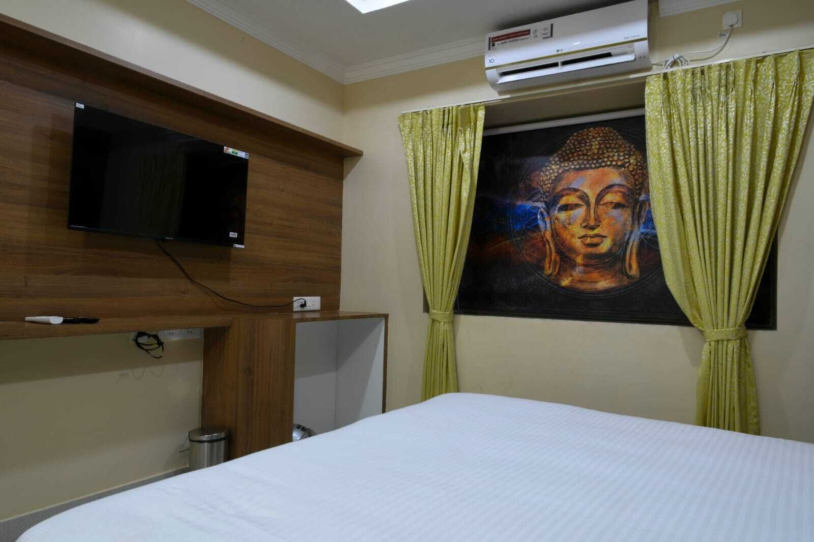 Una habitación de hotel con cama, televisor montado, aire acondicionado y un cuadro de Buda en la pared.