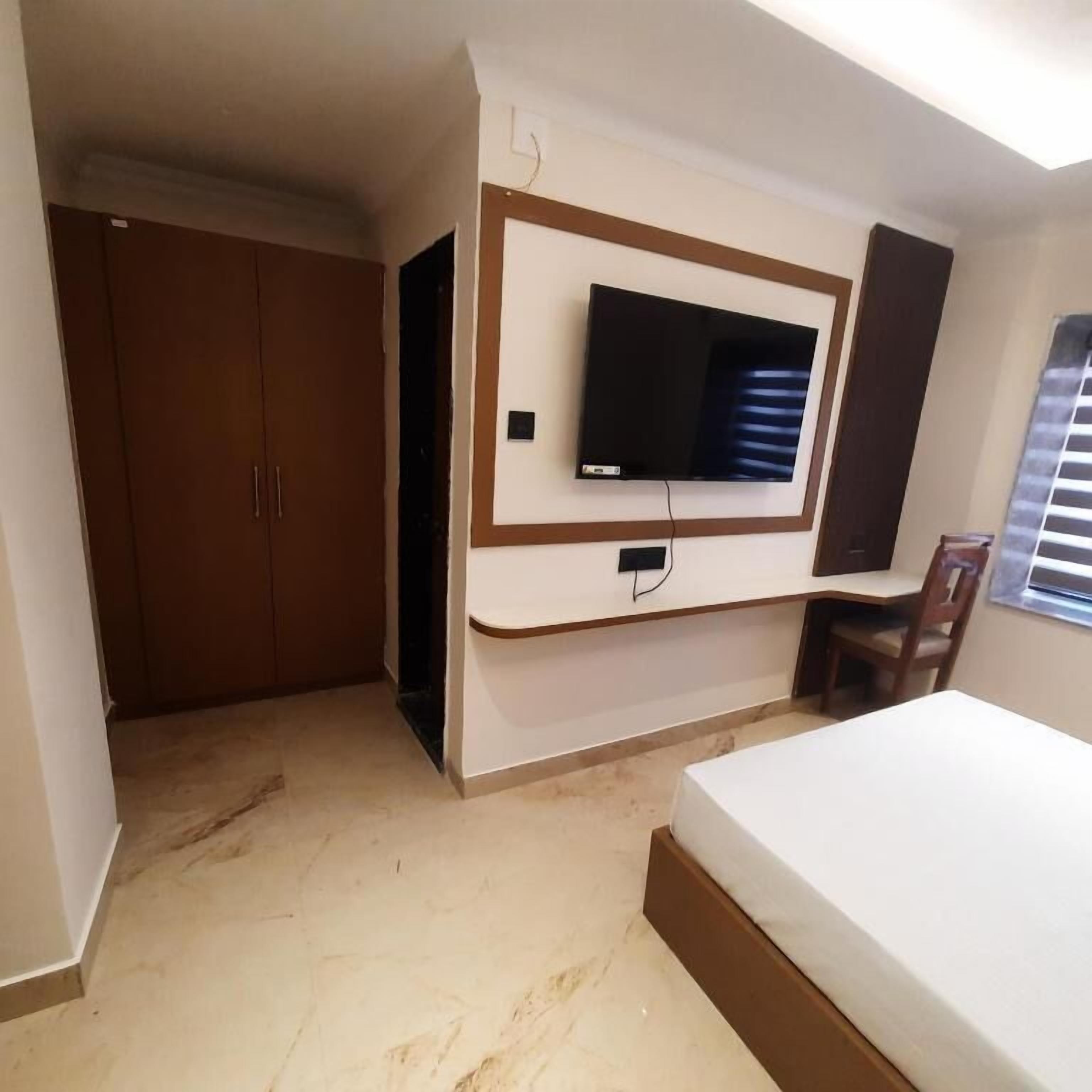 A bedroom with a bed, a TV mounted on the wall, a wardrobe, and a window with blinds.
