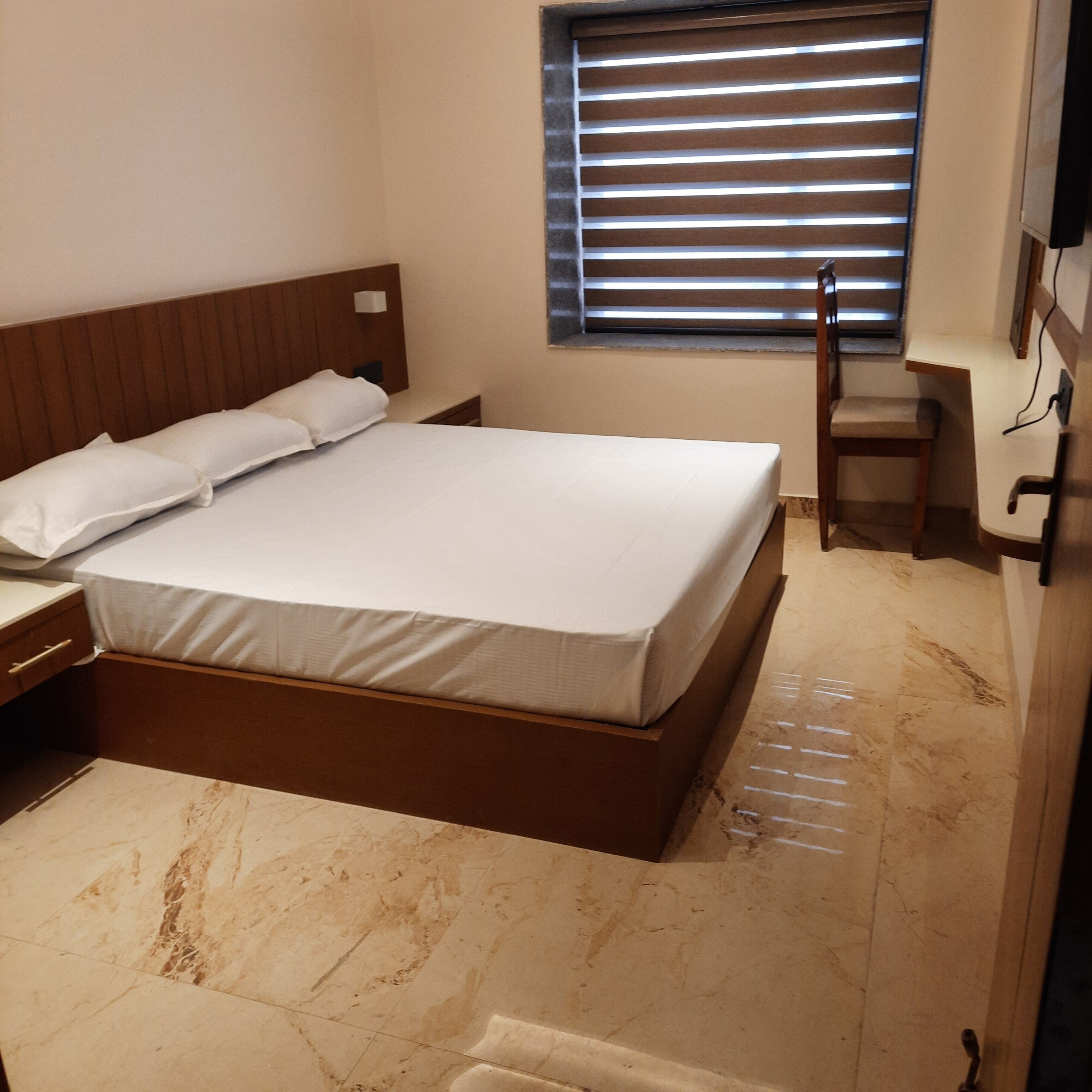 Une chambre d’hôtel avec un grand lit, une tête de lit en bois, une table de chevet, une chaise, une fenêtre avec des stores et un sol en marbre.