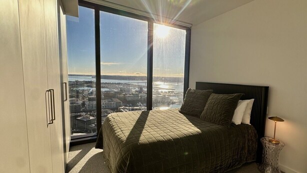 A bedroom with a large bed, a view of a cityscape through a floor-to-ceiling window, and a modern lamp on a side table. - High-Rise Luxury Stay with Harbour Views – 51 Albert Street (Auckland Central)