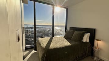 A bedroom with a large bed, a view of a cityscape through a floor-to-ceiling window, and a modern lamp on a side table.