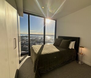 A bedroom with a large bed, a view of a cityscape through a floor-to-ceiling window, and a modern lamp on a side table.