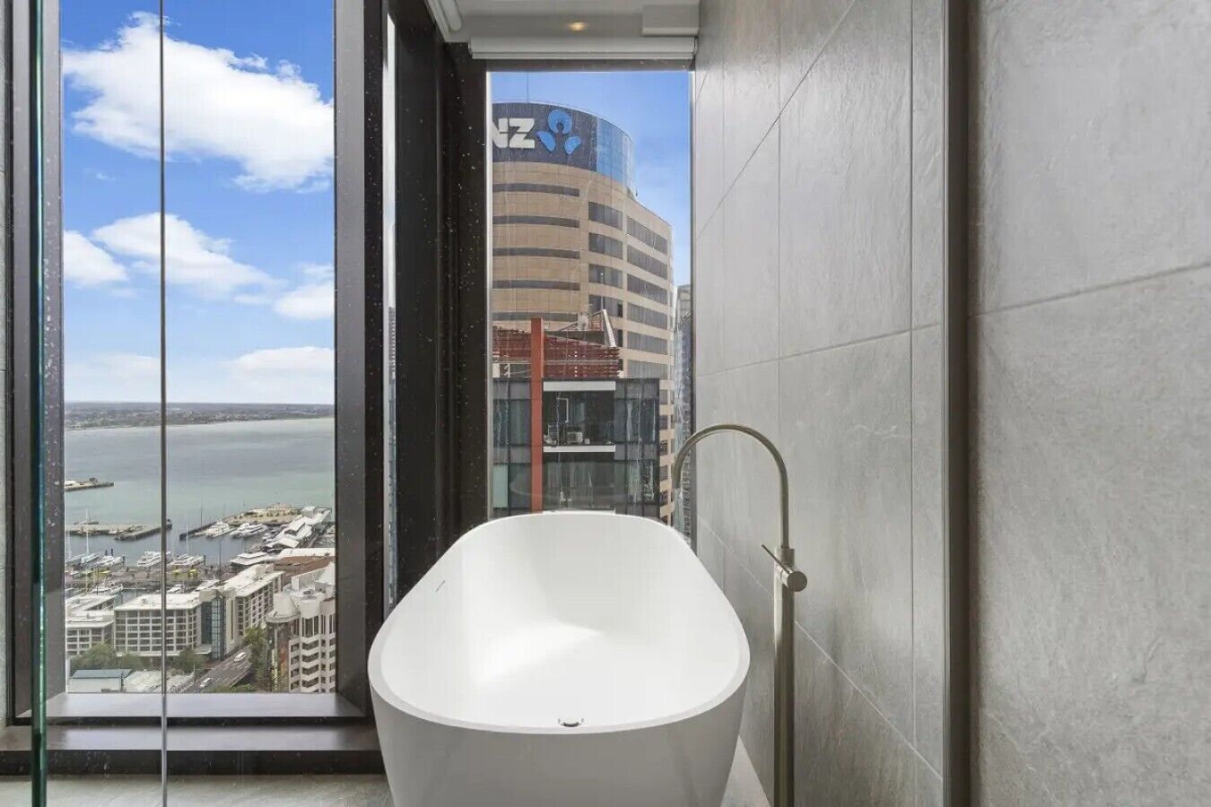 A modern bathroom with a freestanding bathtub, a view of a cityscape through a large window, and a building with a blue logo in the background.