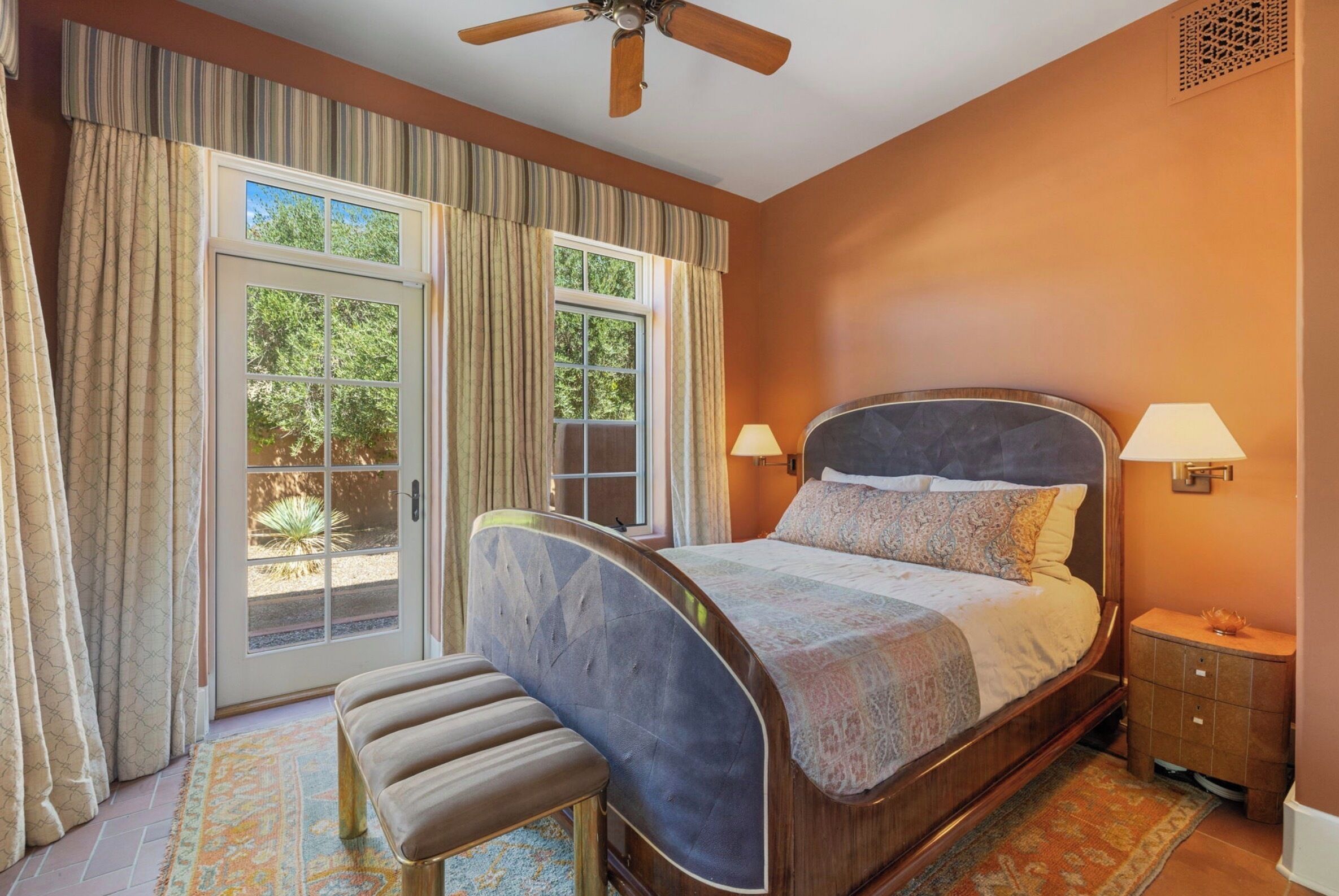A bedroom with a bed, a bench, a nightstand, a lamp, a ceiling fan, and a window with curtains.