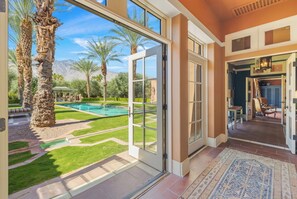 A spacious indoor area with a large glass door leading to a pool and garden, a patterned rug, and a view of palm trees and mountains. - Moroccan Desert Oasis in Serene Neighborhood (Palm Springs)
