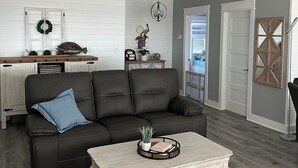 Ein modernes Wohnzimmer mit einem dunkelgrauen Ledersofa, einem Holztisch und einem Deckenventilator, mit Blick auf die Küche im Hintergrund.