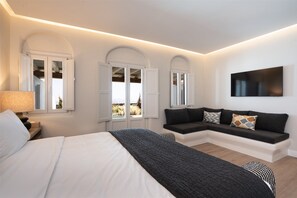 A modern bedroom with a bed, a sofa, a TV, and windows with shutters. - Nero Light-filled Suites (Santorini)