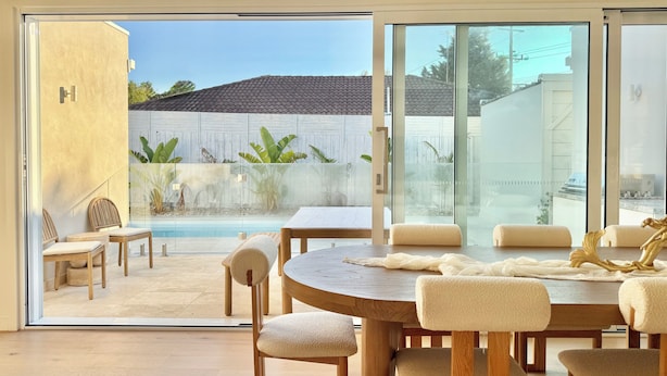 A modern dining area with a glass door leading to a pool, wooden furniture, and a view of a backyard with plants. - Beige Beach House Blairgowrie (Blairgowrie)