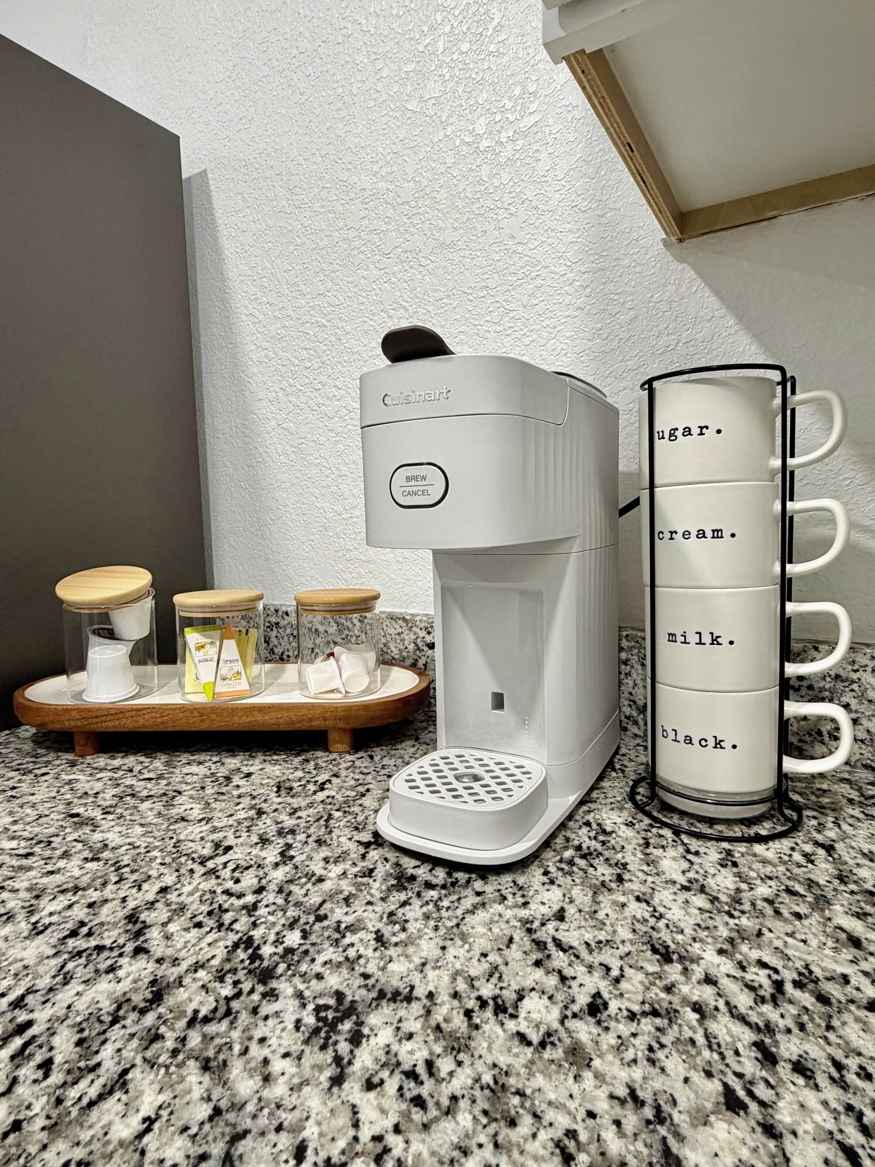 A coffee maker, a mug rack, and a tray with containers on a granite countertop.