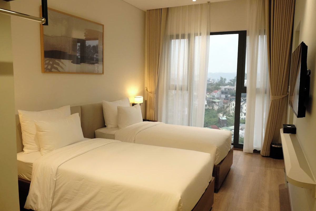 A hotel room with two beds, a large window with curtains, a mounted TV, and a painting on the wall.