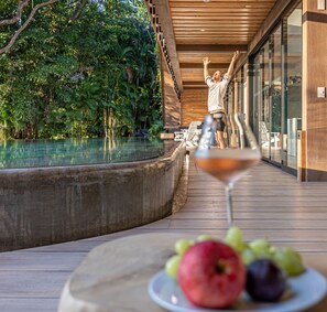Eine Holzterrasse mit Glasgeländer, die auf einen Pool blickt, ein Tisch mit Obst und einem Weinglas, und eine Person, die sich zum Pool streckt.