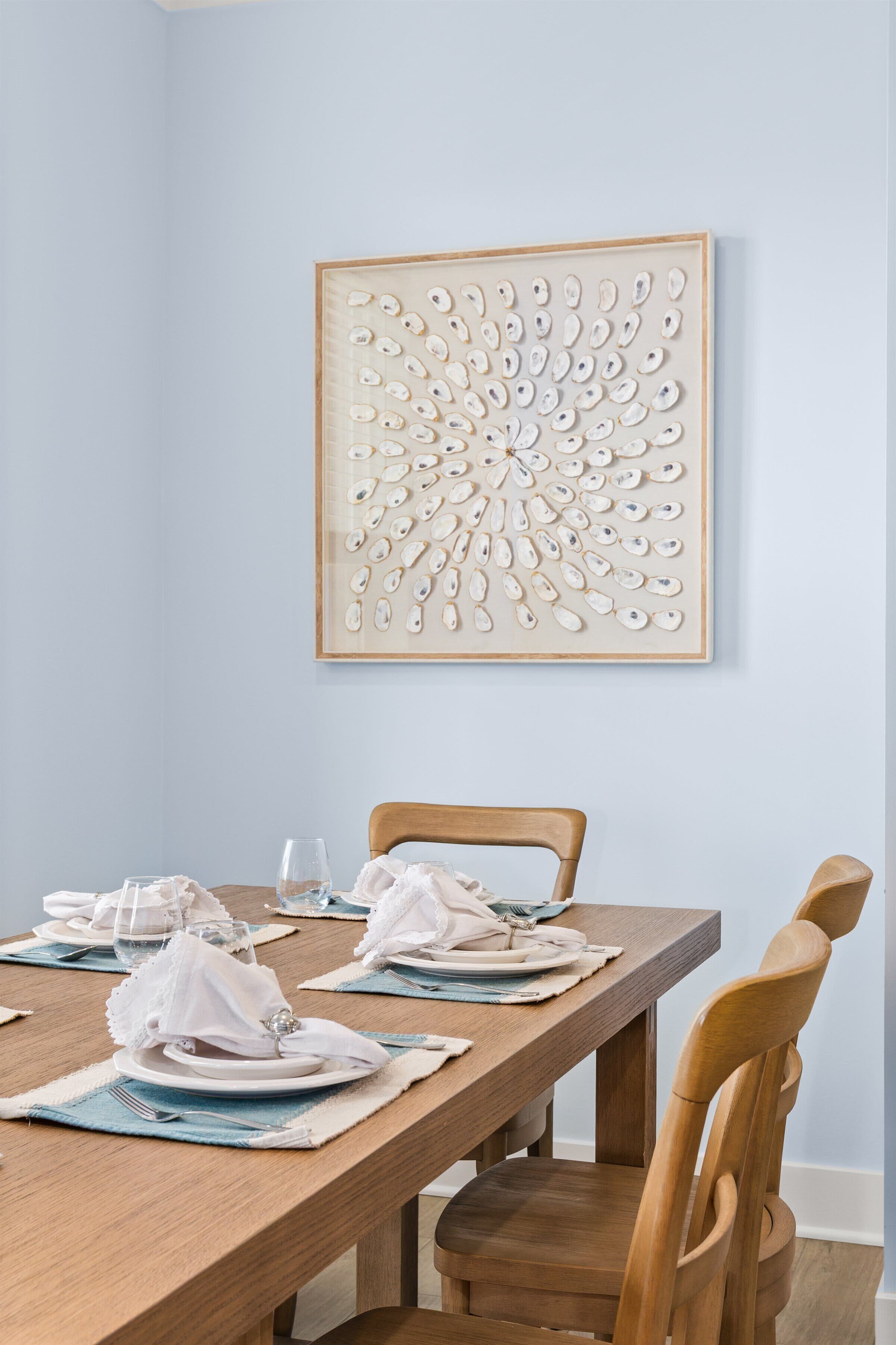 Een eetruimte met een houten tafel gedekt voor twee personen, een lijst met abstracte kunst aan de muur, en een houten stoel.