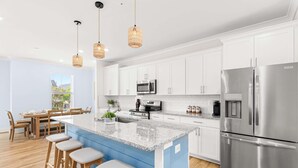 Una cocina moderna con armarios blancos, un refrigerador de acero inoxidable, una isla de granito con taburetes de bar y lámparas colgantes de mimbre.