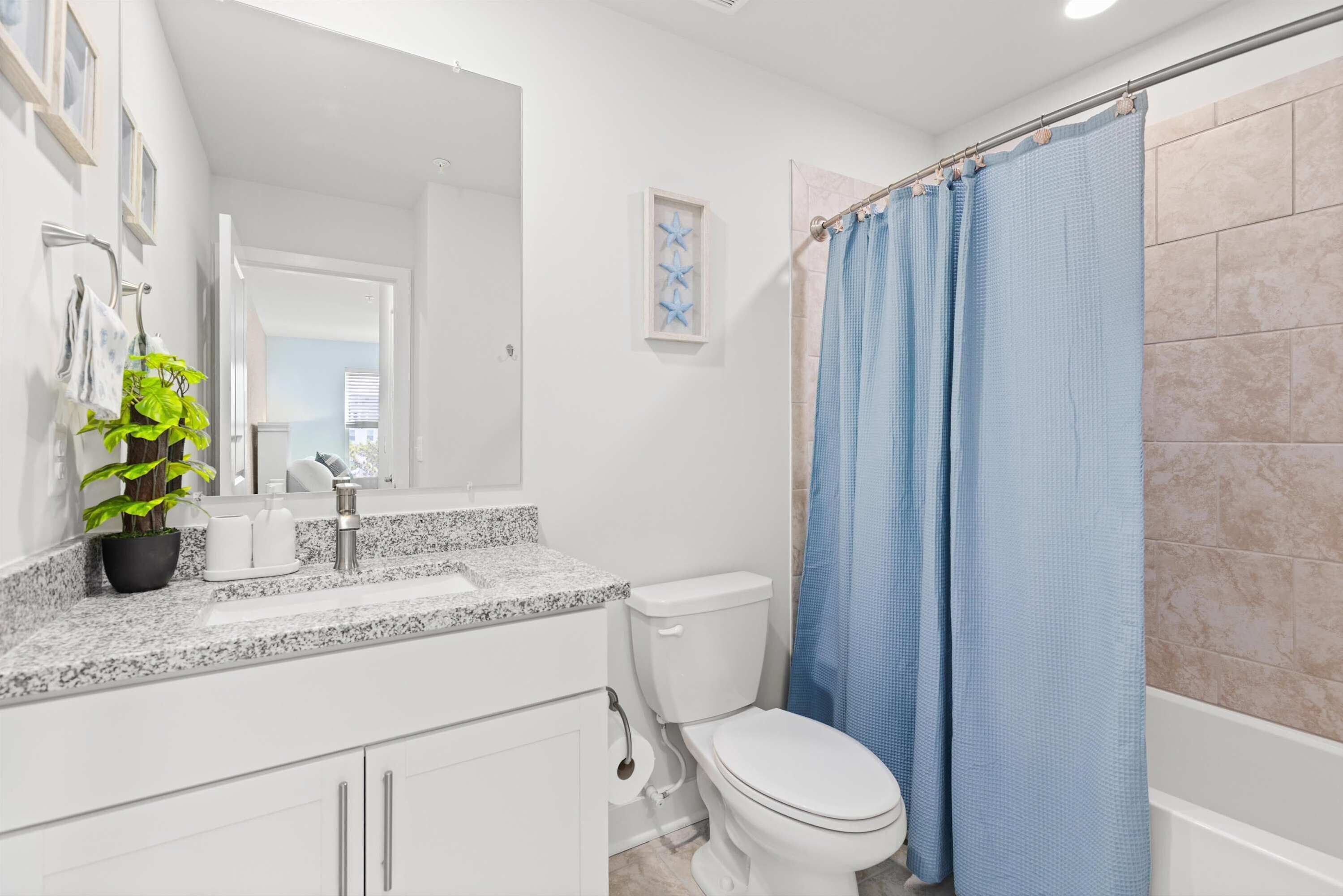 A bathroom with a white toilet, a sink with a granite countertop, a mirror, a potted plant, and a blue shower curtain.