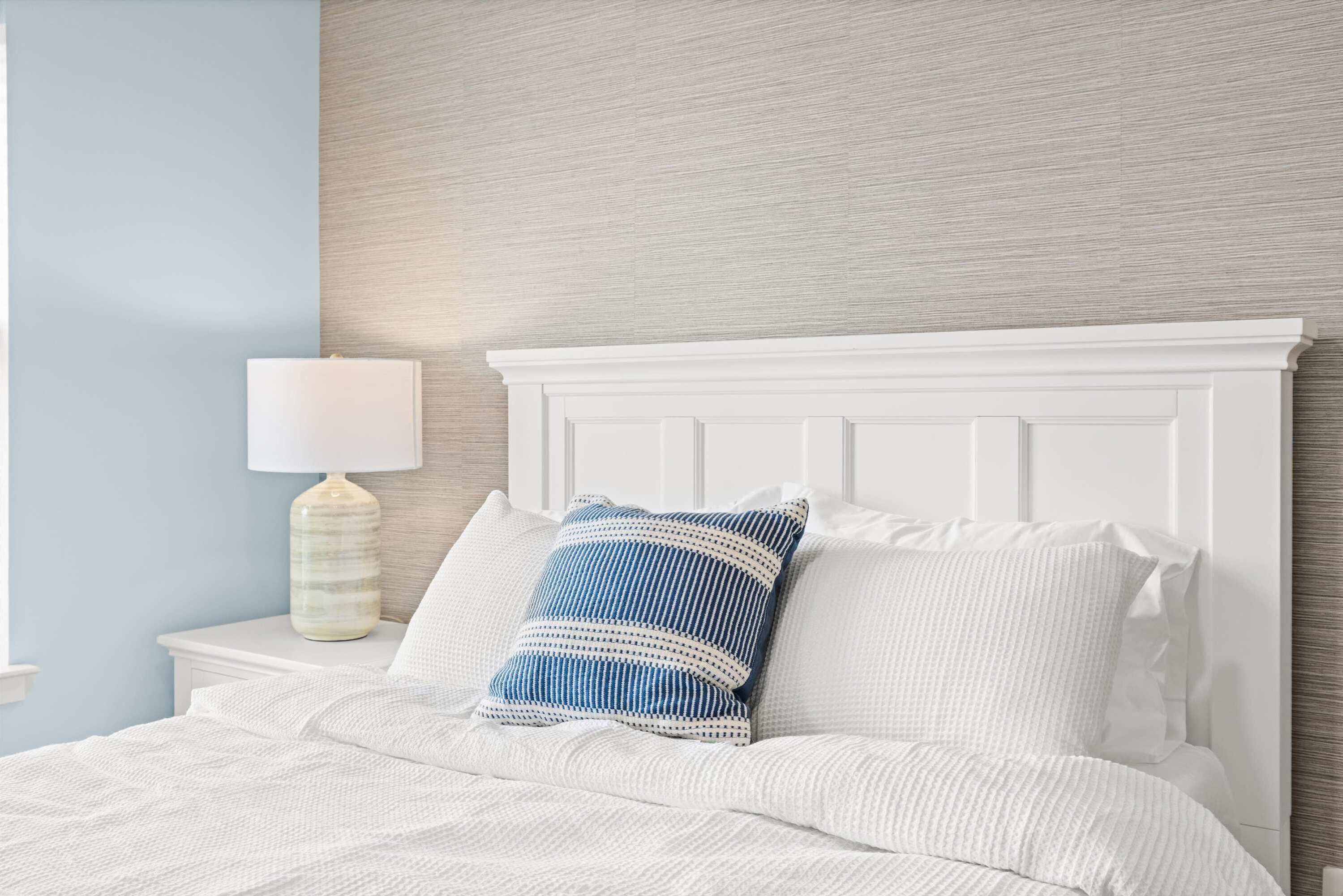 Un lit bien fait, avec une literie blanche et un oreiller à rayures bleues, une table de chevet blanche avec une lampe, et un mur texturé derrière le lit.