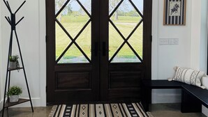 Uma porta de madeira escura com painéis de vidro em padrão de losangos, um porta-roupas de metal preto, um quadro na parede e um banco com um tapete estampado.