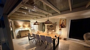 A modern dining area with a wooden table and chairs, pendant lights, and a kitchen in the background. - 5-bedroom house, sea view, swimming pool (Fréjus)