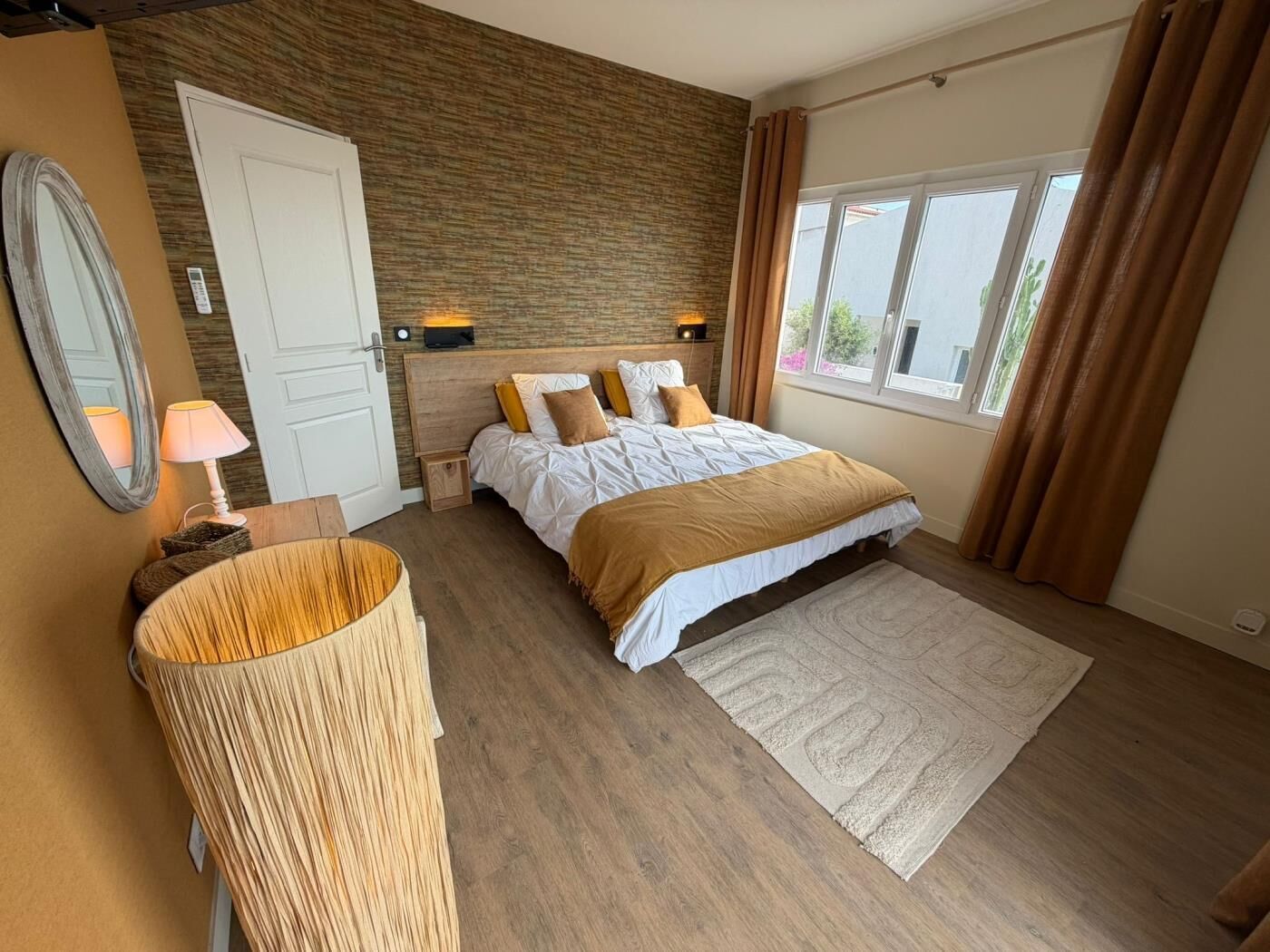Een slaapkamer met een bed, nachtkastje, spiegel, lamp, vloerkleed en een raam met gordijnen.