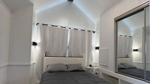 Een slaapkamer met een bed, nachtkastje, spiegel en raam met gordijnen.